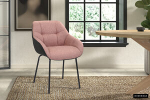 Het SALE - Eetkamerstoel 'Sem' Stof Roze Vloerkleed PU Donkergrijs geeft een modern tintje aan een lichte kamer met zijn roze bekleding en zwarte poten, geplaatst op een vloerkleed met structuur bij een ronde houten tafel en een groot raam.