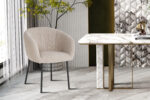 Een paar SALE - Eetkamerstoel 'Sevilla' Bouclé Grijs stoelen staat naast een marmeren tafel met gouden accenten in een stijlvolle kamer met gestructureerde muren, een marmeren vloer en een plant in pot.
