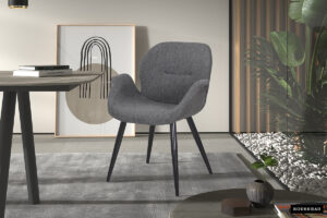 De SALE - Stoel 'Maxim' Stof Grijs (2 per doos) is een moderne grijs gestoffeerde stoel met zwarte poten, getoond in een hedendaagse kamer met een grijs vloerkleed, ronde tafel, abstracte muurkunst, kamerplanten en een rotsbed.