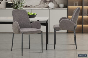 Twee Eetkamerstoel 'Bilbao' Bouclé Grijs stoelen (set van 2) met gebogen armleuningen en metalen poten staan in een stijlvolle keuken met een marmeren achterwand, aanrecht en planken met groene appels. Het ROUSSEAU-logo verschijnt in de hoek.