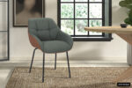 De SALE - Stoel 'Sem' heeft een groene stoffen bekleding en cognac PU zijkanten, hier getoond op een geweven vloerkleed in een lichte kamer met een groot raam, houten tafel en muurkunst. Verkocht als set van 2 per doos.
