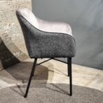 De Eetkamerstoel Malaga heeft een gebogen rugleuning, zachtgrijze bekleding en slanke zwarte metalen poten. Hij staat op een houten vloer met een rond vloerkleed gedeeltelijk zichtbaar eronder.