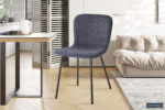 In een moderne kamer staat de Stoel 'Susan' Bouclé Donkergrijs (set van 2) met zwarte metalen poten naast een houten tafel, een lichtbeige bank en een vloerkleed met patroon op een houten vloer met visgraatmotief. Zonlicht verlicht de gezellige ruimte.