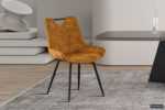 In een moderne eetkamer staat de 'Nina' Eetkamerstoel Fluweel Goud - mosterdgele fluwelen stoelen met zwarte metalen poten - op een grijs vloerkleed, in combinatie met een strakke tafel en een minimalistische, neutraal getinte inrichting.