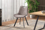 De Stoel 'Pablo' Micro fiber PU Grijs (2 per doos) is een moderne taupe gestoffeerde stoel met zwarte metalen poten, hier gezien op een vloerkleed met patroon naast een houten vloer, een houten bureau, speakers, planten en hoge ramen met transparante gordijnen.