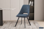 Het SALE - Eetkamerstoel 'Filou' Bouclé Blauw Vloerkleed PU Blauw (2 per doos) heeft een modern blauw gestoffeerd ontwerp met zwarte schuine poten, geplaatst op een beige getextureerd vloerkleed in een minimalistische kamer met lichte kasten en een marmeren tafelrand.