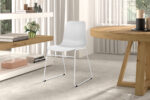 In een moderne, minimalistische kamer staan Stoel 'Marie' PP Wit stoelen (2 per doos) op een lichtgrijze vloer, in combinatie met een ronde houten tafel, een plank met boeken en bloemen en grote ramen met witte gordijnen die natuurlijk licht binnenlaten.