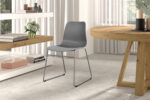 Een moderne eetkamer met Stoel 'Juta' Fluweel Roze stoelen (2 per doos) op metalen poten, een ronde houten tafel, planken met boeken, vazen en bloemen. Grote ramen met transparante gordijnen laten natuurlijk licht binnen.