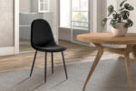 De Stoel 'Boy' Fluweel Zwart (2 per doos) staat op een vloerkleed met structuur naast een ronde houten tafel met daarop een potplant, in een lichte, minimalistische ruimte met grote ramen en lichtgekleurde muren.