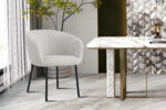 Een modern interieur met Stoel 'Sevilla' Stof Beige stoelen en zwarte poten naast een wit marmeren tafel met gouden accenten; de achtergrond heeft gestructureerde muren, een bladplant en grote ramen.