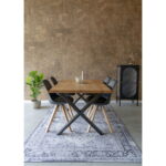 Een moderne eethoek met een houten tafel, vier Bergen Eetkamerstoelen - zwarte stoelen met poten van natuurlijk hout - een blauw vloerkleed met patroon, een potplant en een klein zwart kastje tegen een bruine muur met structuur.