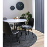 Een moderne eethoek met een ronde witte tafel, twee sets Stockholm eetkamerstoelen in zwart PU met zwarte poten (in totaal vier stoelen), een zwart cirkelvormig vloerkleed, twee wandklokken, planten in potten, kaarsen en verticale grijze jaloezieën voor het raam.