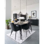 Moderne eetkamer met twee sets Harbo Dining Chairs in zwart PU met zwarte poten rond een zwarte tafel op een vloerkleed met patroon. Erboven hangen drie hanglampen, met planten, kasten, muurkunst, neutrale muren en een grijze tegelvloer.