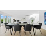 Moderne eetkamer met een grote zwarte tafel en Harbo Dining Chairs in donkergrijs PU met zwarte poten (set van 2) op een lichthouten vloer, witte muren, grote ramen met groen en een minimalistische inrichting met planten en kunstwerken.