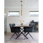 Moderne eetkamer met een houten tafel, zes Harbo draaibare eetkamerstoelen in zwart PU en zwarte poten (HN1223), hanglamp, vazen met gedroogde planten, twee ramen voor natuurlijk licht, een vloerkleed met patroon onder de tafel en een kast in de hoek.