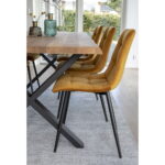 Een houten eettafel met zwarte metalen poten wordt omringd door Middelfart Dining Chairs in mosterdgeel fluweel met zwarte poten (set van 2), allemaal geplaatst op een lichtgrijs vloerkleed in een lichte kamer met grote ramen.