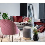 Een moderne woonkamer met een grijze bank, rode en roze fluwelen kussens, een Geneve eetkamerstoel van roze fluweel met poten in messinglook, een grijze voetenbank, een ronde zwarte tafel met kaarsen en vetplanten, een zwarte vaas met planten en een grote vloerspiegel.