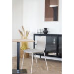 Een moderne eethoek met een lichtgrijze Dining Chair HN1263 aan een houten tafel, gedroogde planten in een rieten mand, een zwarte kast met glazen deuren en abstracte kunst op een witte muur.