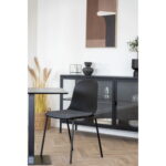 De Dining chair, zwart, HN1264 (set van 2) met slanke zwarte poten en zitting staat bij een bijpassende zwarte tafel voor een kast met glazen voorkant, geaccentueerd door decoratieve vazen, abstracte kunst en gedroogd gras voor een minimalistische look.