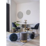 Een moderne eethoek met een ronde witte tafel, vier Geneve stoelen van grijs fluweel met zwarte poten, een kleine grijze voetenbank, een glazen salontafel, planten in potten en twee ronde spiegels op een witte muur boven een vloerkleed met patroon.