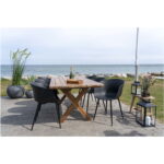 Een houten eettafel voor buiten met drie sets Roda Dining Chairs in zwart staat opgesteld op een patio aan zee, met groene planten, kaarslantaarns en champagne, met een grasveld en de oceaan erachter.