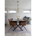 Moderne eetkamer met een rechthoekige houten tafel, zes Harbo draaibare eetkamerstoelen in vintage bruin PU en zwarte poten, een vloerkleed met patroon, minimalistische hanglamp, decoratieve vazen met gedroogde planten en twee horizontale ramen voor natuurlijk licht.