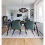 Moderne eetkamer met een zwarte tafel, twee sets Harbo Dining Chairs in groen fluweel, een geweven hanglamp en lichte houten vloeren. Grote ramen bieden natuurlijk licht, terwijl planken met decor en een houtkachel de ruimte compleet maken.