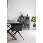 Minimalistische eetkamer met een houten tafel, Harbo draaibare eetkamerstoel in donkergrijs PU en zwarte poten (HN1221), een glazen kast voor glaswerk, decoratieve vazen met gedroogde planten en een groene potplant in de hoek tegen een witte muur.