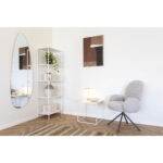 Een moderne, minimalistische kamer met de grijze Lana eetkamerstoel, een kleine witte bijzettafel met een lamp, een wit rek met decor, een grote ovale spiegel, abstracte muurkunst en een vloerkleed met patroon op een houten vloer.
