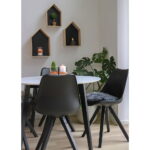 Een moderne eethoek met Bergen Eetkamerstoel stoelen in zwart met zwarte houten poten rond een witte ronde tafel, geaccentueerd door een monstera in pot, kaarsen en huisvormige wandplanken op een witte muur.
