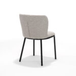 De Kim stoel Stof Teddy Natural is een moderne stoel met een licht naturelkleurig gestoffeerde zitting en rugleuning, slanke zwarte metalen poten, afgebeeld onder een hoek om de rugleuning en zijkant te tonen tegen een effen witte achtergrond.