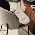 De Eetkamerstoel Lio - Naturel is een moderne stoel met een beige stoffen zitting en rugleuning, zwarte metalen poten en staat naast een ronde houten tafel in een zonovergoten kamer.