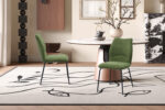 Een moderne eethoek met twee Stoel 'Lara' Stof Ote Sea 360 Groen stoelen, een ronde wit marmeren tafel met een gouden onderstel, een decoratieve vaas met takken en een abstract vloerkleed met zwarte lijntekeningen op een lichte achtergrond.