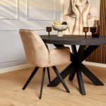 Een moderne eethoek met een ronde zwarte tafel, Eetkamerstoel Nova Beige, bruin glaswerk, een fruitschaal, grote muurschilderingen van een vrouw in zachte tinten, houten vloeren en gedeeltelijk houten lambrisering.