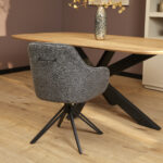 Een moderne eetkamer toont de Eetkamerstoel Jens in antraciet, met een gebogen rug en zwarte metalen poten, in combinatie met een houten tafel met een zwart X-onderstel. Een decoratieve vaas met gedroogde takken siert het tafelblad.