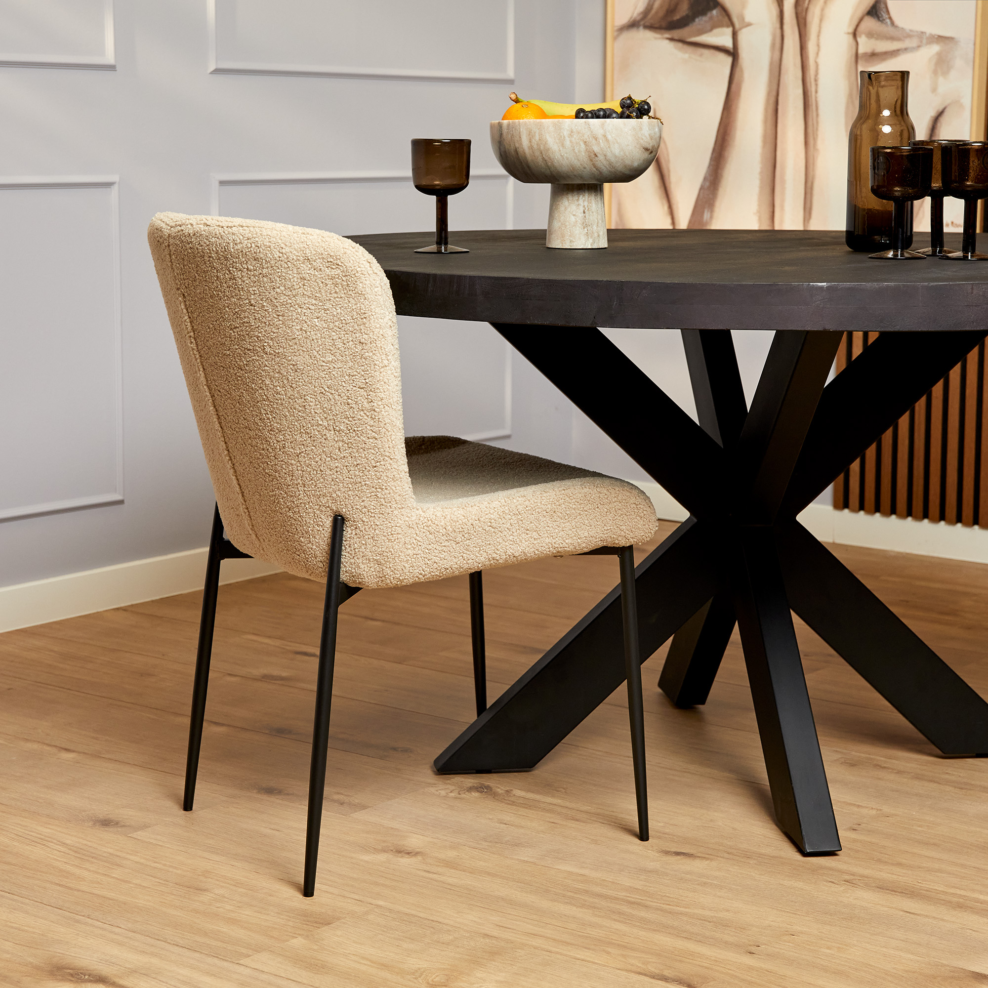 Een moderne eetkamer met een ronde zwarte tafel, de Eetkamerstoel Tess Beige met zwarte poten, decoratief glaswerk, een stenen fruitschaal, abstracte muurkunst en een lichte houten vloer.