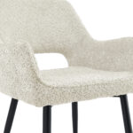 Een close-up van de Eetkamerstoel Ray - Beige, met beige bekleding en zwarte schuine poten. Slechts een deel van de zitting, rugleuning en armleuningen is zichtbaar.