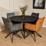 Een ronde zwarte tafel met vier fluwelen stoelen, waaronder de Eetkamerstoel Dani Velvet Hunter Green, staat op een houten vloer. Op tafel staat een vaas met kleurrijke bloemen en aan de muur hangt ingelijste kunst.