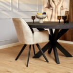 Een moderne eethoek met de Eetkamerstoel Nova Champagne, een ronde zwarte sculpturale tafel, bruin getinte glazen, een fruitschaal, houten vloeren en een kunstwerk van een vrouw aan de muur.