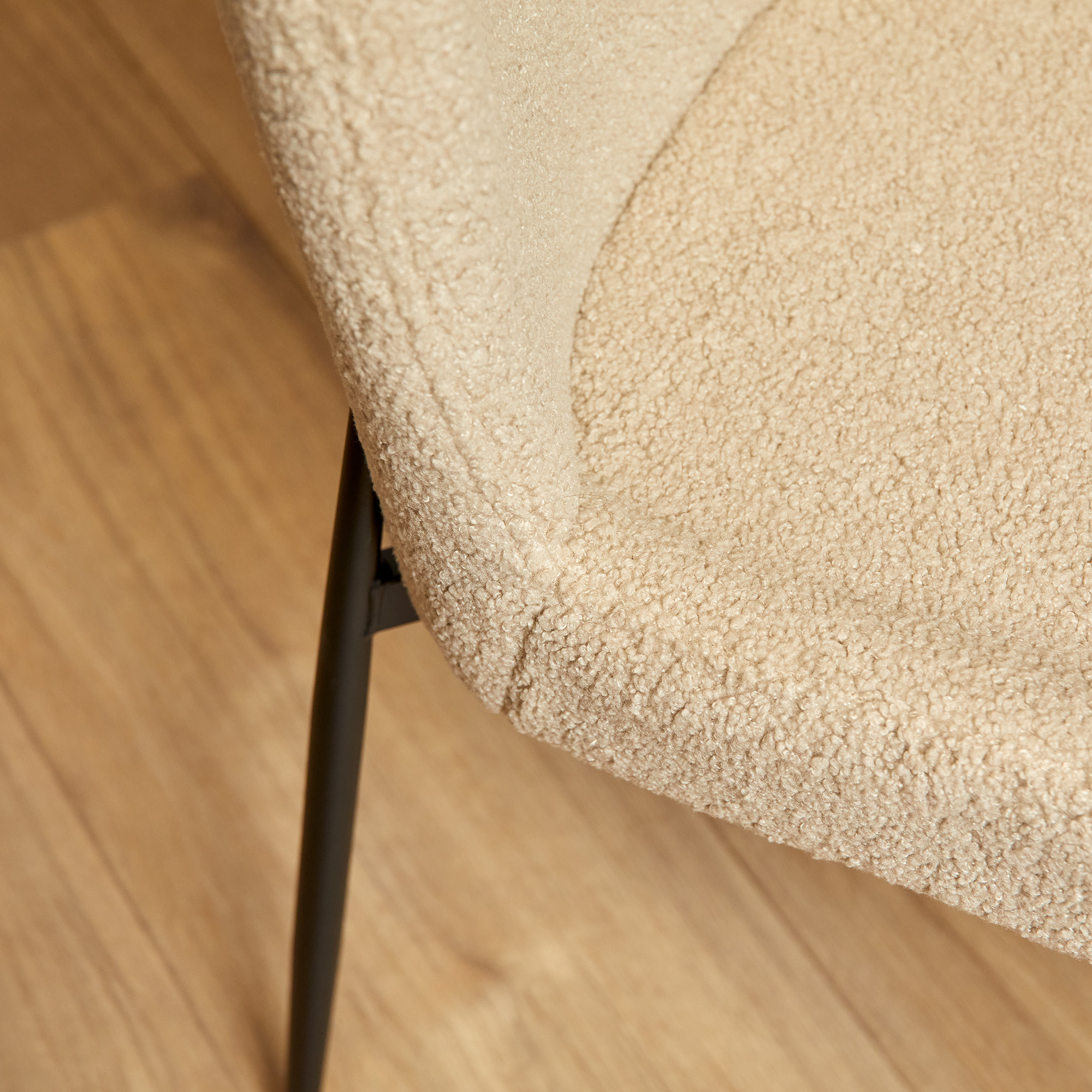 Close-up van de Eetkamerstoel Tess Beige, met zachte, gestructureerde bekleding en zwarte metalen poten, getoond op een houten vloer. De afbeelding benadrukt de knusse stof en het moderne ontwerp.