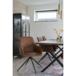 Een moderne eetkamer met Harbo Dining Chair with Swivel in vintage bruin PU en zwarte poten, een houten tafel met zwarte metalen poten, een vloerkleed met patroon, glazen kast, hanglampen en droogbloemen op tafel bij een zonovergoten raam.
