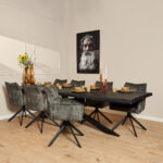 Een moderne eetkamer met een zwarte rechthoekige tafel, zes Eetkamerstoel Dani fluwelen jagergroene stoelen, taankleurig servies, hoge bloemenvazen, kaarsen en een muurportret van een oudere man met een lange grijze baard op beige muren en lichthouten vloeren.