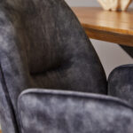 Close-up van de Eetkamerstoel Nova Antraciet naast een houten tafel, met de zachte donkergrijze bekleding en de elegante gebogen rugleuning. Een vaag decoratief object verschijnt op de achtergrond.