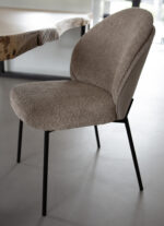 Een set van twee taupe/beige eetkamerstoelen zonder armleuningen, met gebogen gestructureerde zittingen en zwarte metalen poten, staan naast een houten tafel met natuurlijke randen op een gladde lichtgrijze vloer.