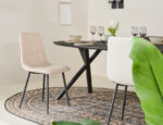 Een moderne eethoek heeft een zwarte tafel voor vier personen, Eetkamerstoel Yerevan - Bouclé Taupe stoelen, een vloerkleed met ovaal patroon, groene plantenbladeren op de voorgrond, neutrale muren en ingelijste kunst voor een minimalistisch tintje.