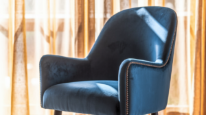 Een fauteuil van blauw fluweel met gebogen armleuningen en decoratieve studs, geïnspireerd op blauwe eetkamerstoelen, staat voor transparante gordijnen in het zonlicht.