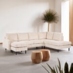 Een lichte, moderne woonkamer met de RIB460 beige 3-zits hoekbank rechts (stof Lincoln 03), twee houten zeshoekige salontafels, een hoge plant in de hoek, een minimale inrichting en neutrale muren en vloeren.