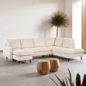 Een lichte, moderne woonkamer met de RIB460 beige 3-zits hoekbank rechts (stof Lincoln 03), twee houten zeshoekige salontafels, een hoge plant in de hoek, een minimale inrichting en neutrale muren en vloeren.