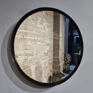 De Spiegel Antiek Brons 66, een ronde spiegel op een grijze muur, weerspiegelt een verweerd oppervlak en een tafeltje met een donkere vaas met gedroogde takken, terwijl op de achtergrond een plant en een gang verschijnen.