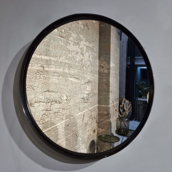 De Spiegel Antiek Brons 66, een ronde spiegel op een grijze muur, weerspiegelt een verweerd oppervlak en een tafeltje met een donkere vaas met gedroogde takken, terwijl op de achtergrond een plant en een gang verschijnen.